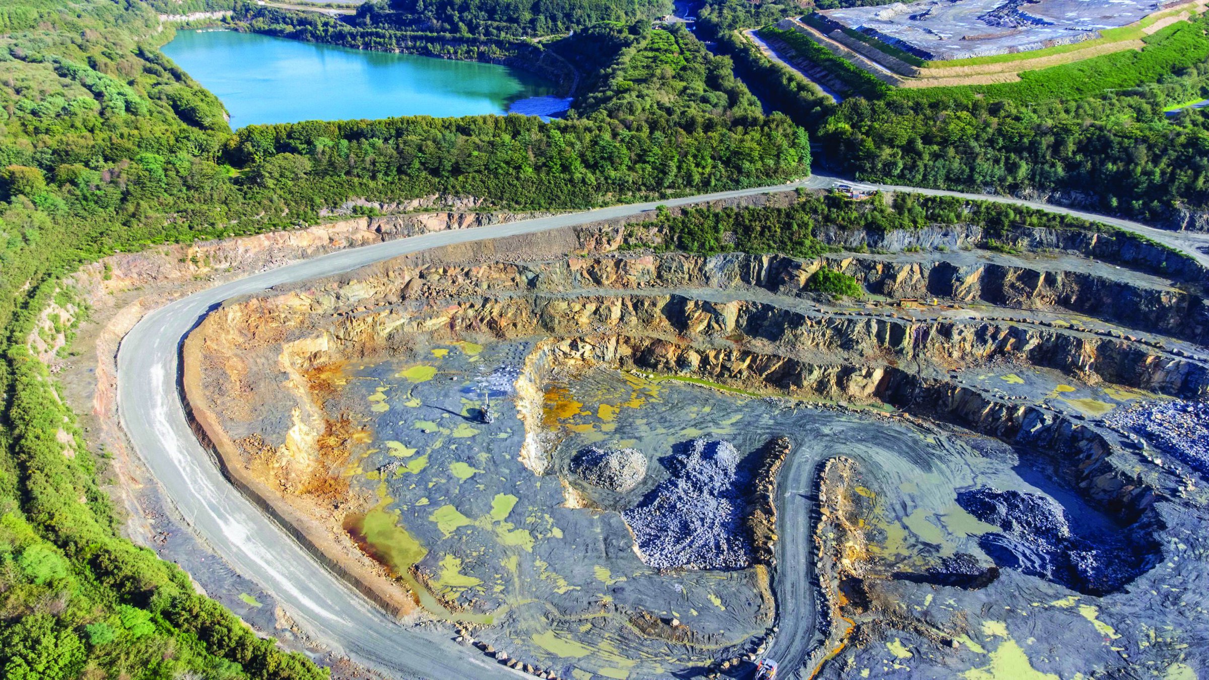 Open-pit quarry with green forest surroundings and water bodies, extracting minerals for ceramics production.