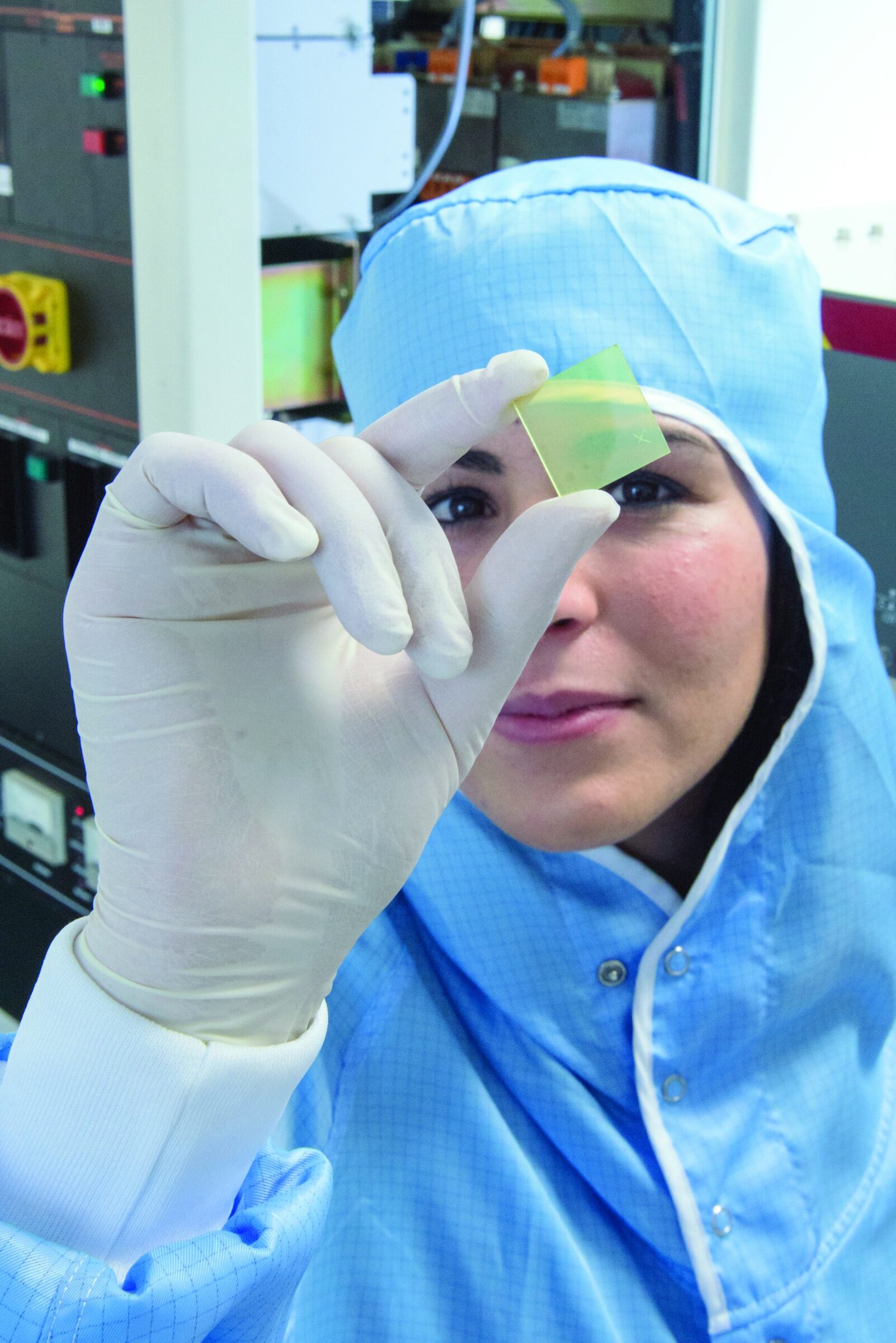 Woman scientist dressed in lab gear holding up a yellow square (oxide thin film).