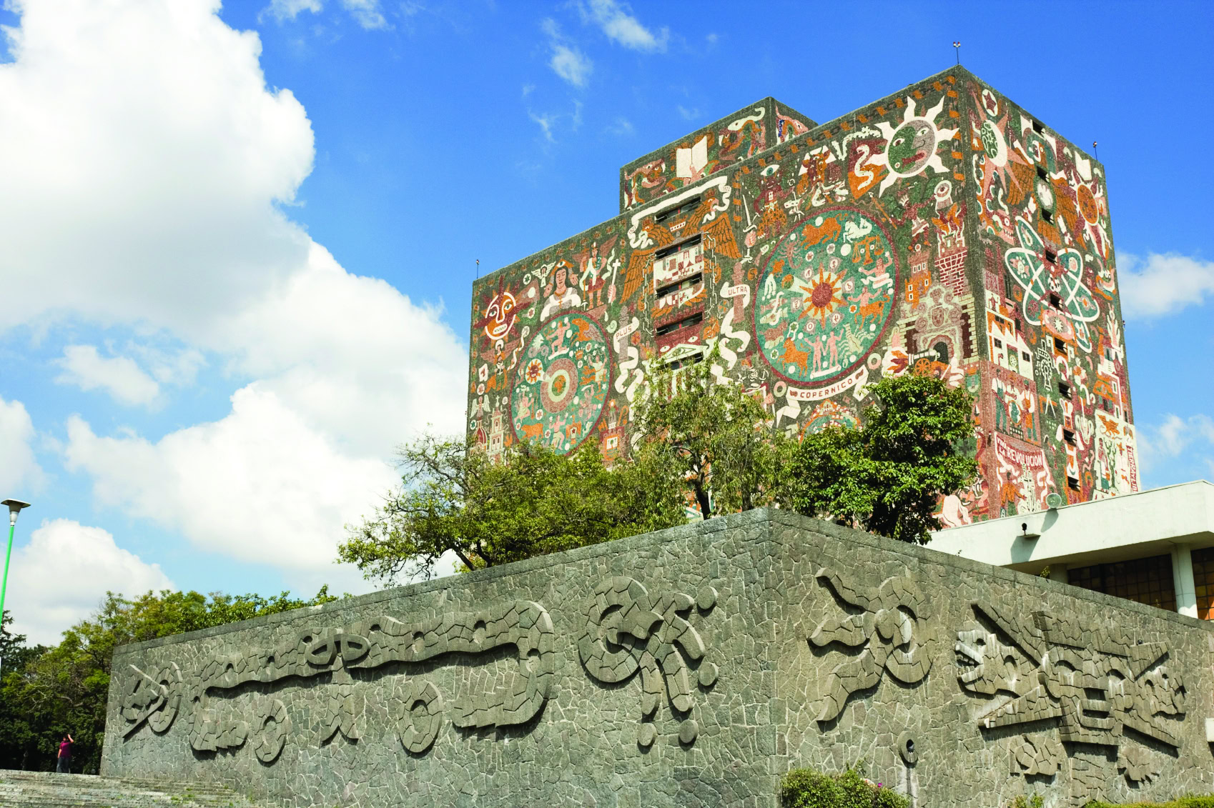 Colorful mural on a building facade with symbolic designs and greenery, under a blue sky with clouds.
