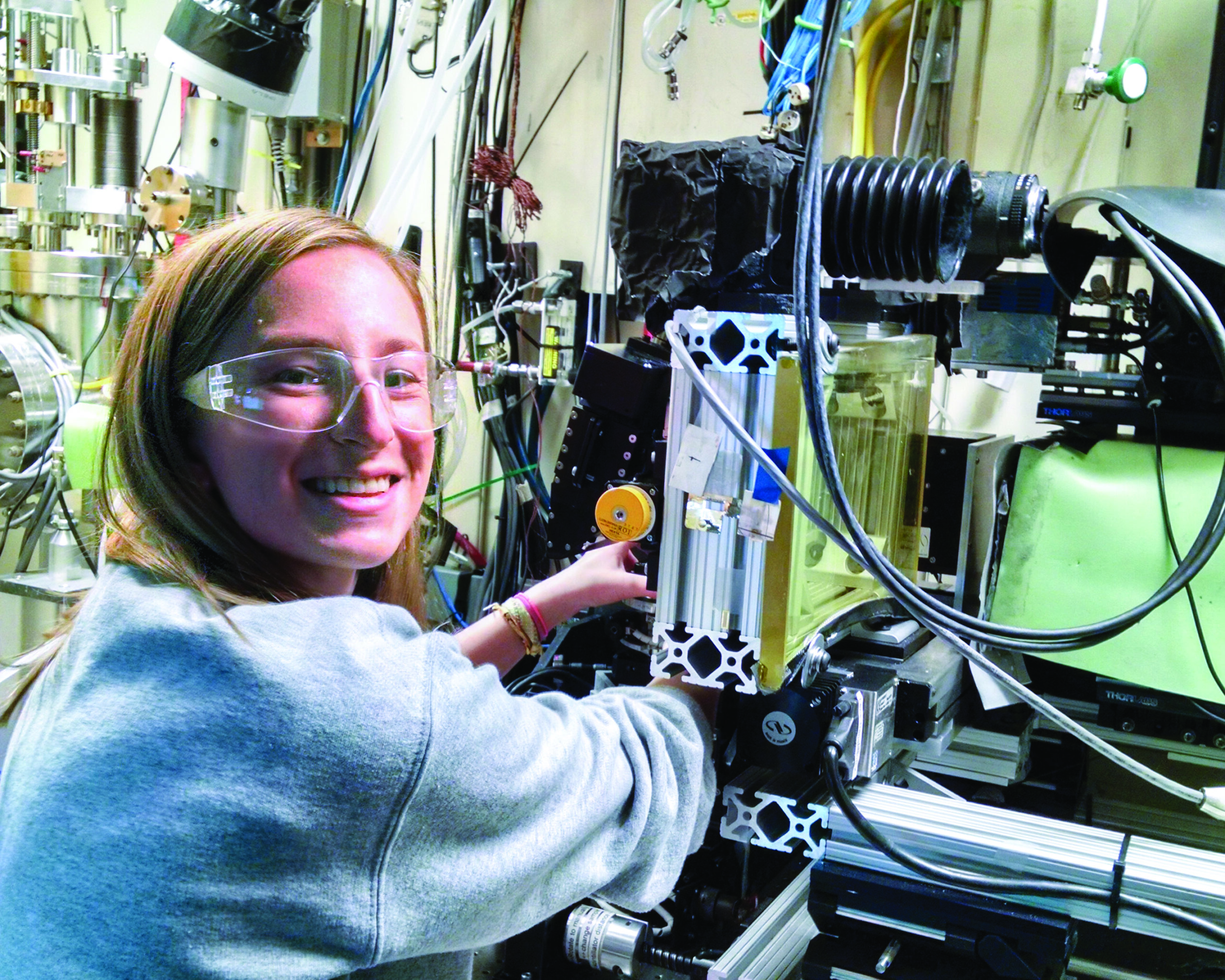 Blond female student smiling in front of x-ray computing machinery in lab setting.