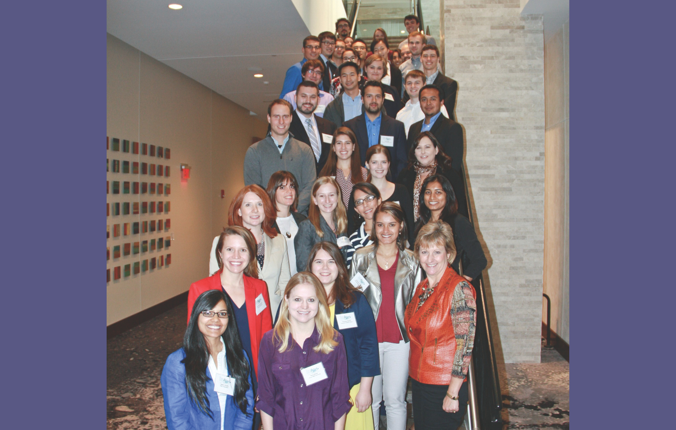 Group of diverse students pose on staircase with purple background behind the photo.