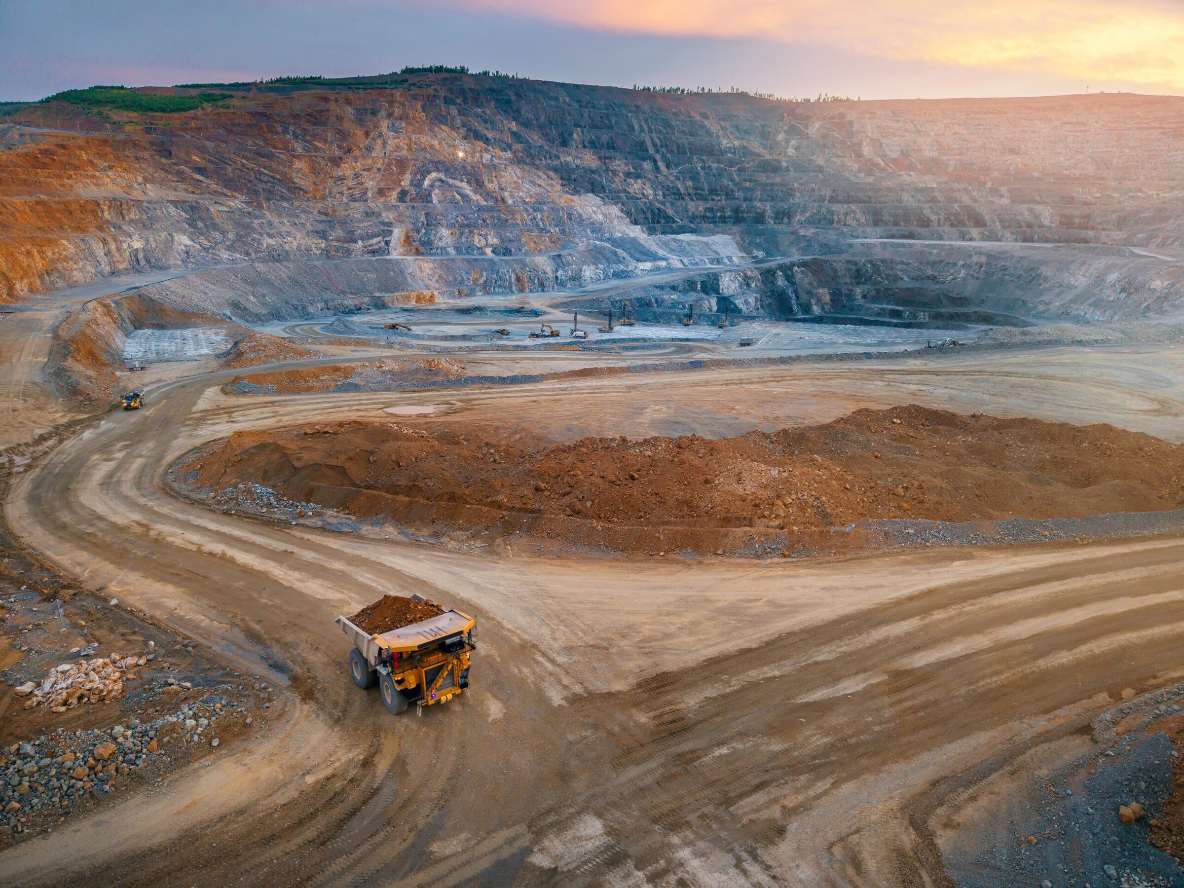 Open-pit mining site with a large dump truck on a dirt road, surrounded by terraced excavations and rocky terrain at sunset.