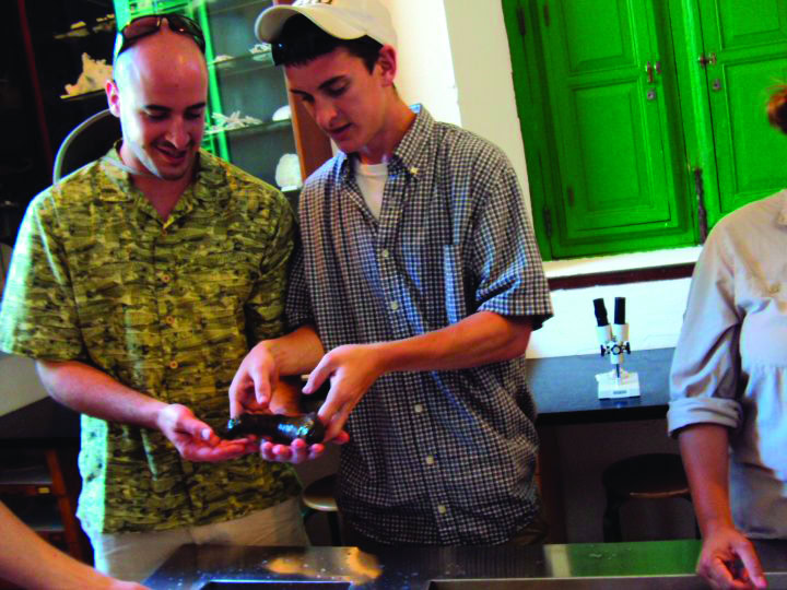 Two young men examine a small object together in a room with green cabinets and scientific equipment.