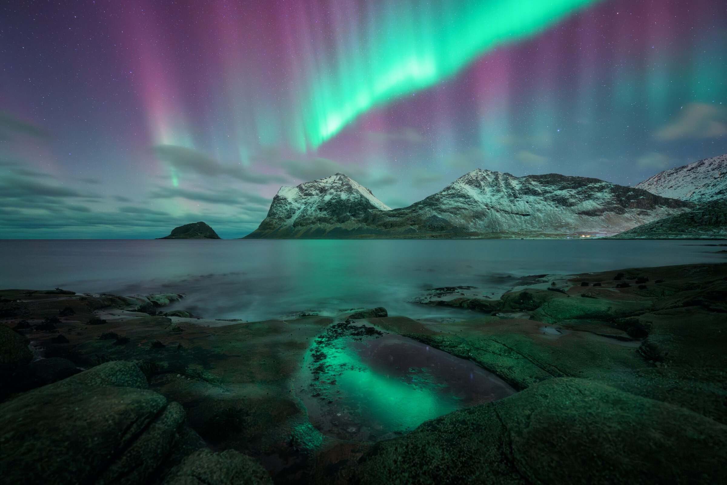 Northern lights over snow-capped mountains and a rocky shoreline at night.