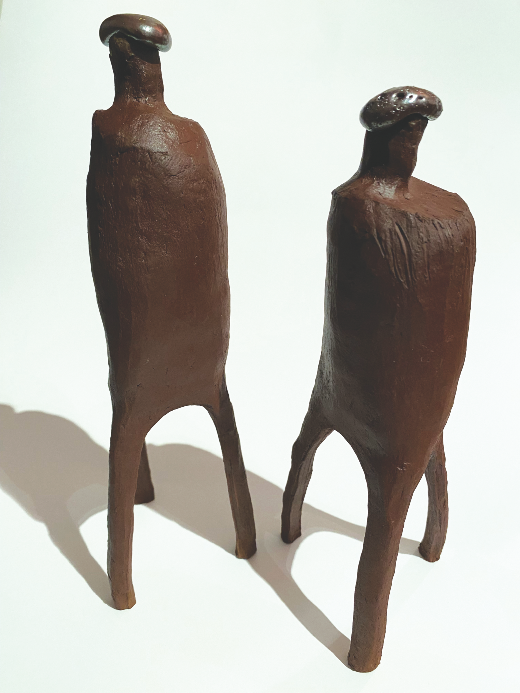 Two abstract ceramic sculptures resembling elongated animals with four legs, dark brown in color, casting shadows on a white background.