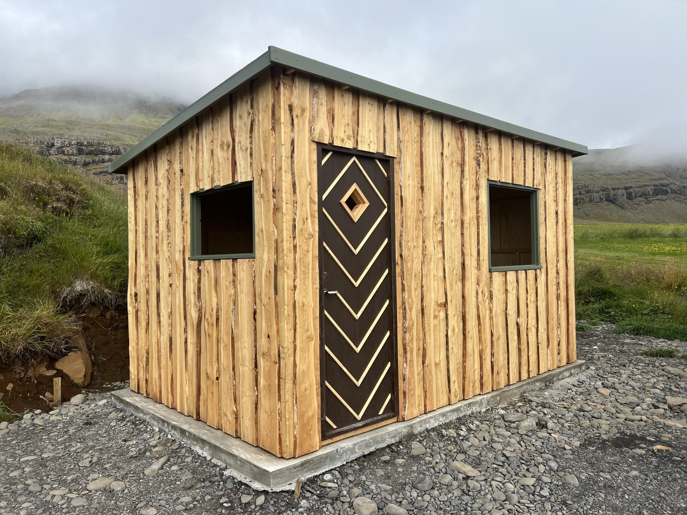 Small wooden cabin with geometric patterned door, set in a grassy landscape with mountains and cloudy sky in the background.