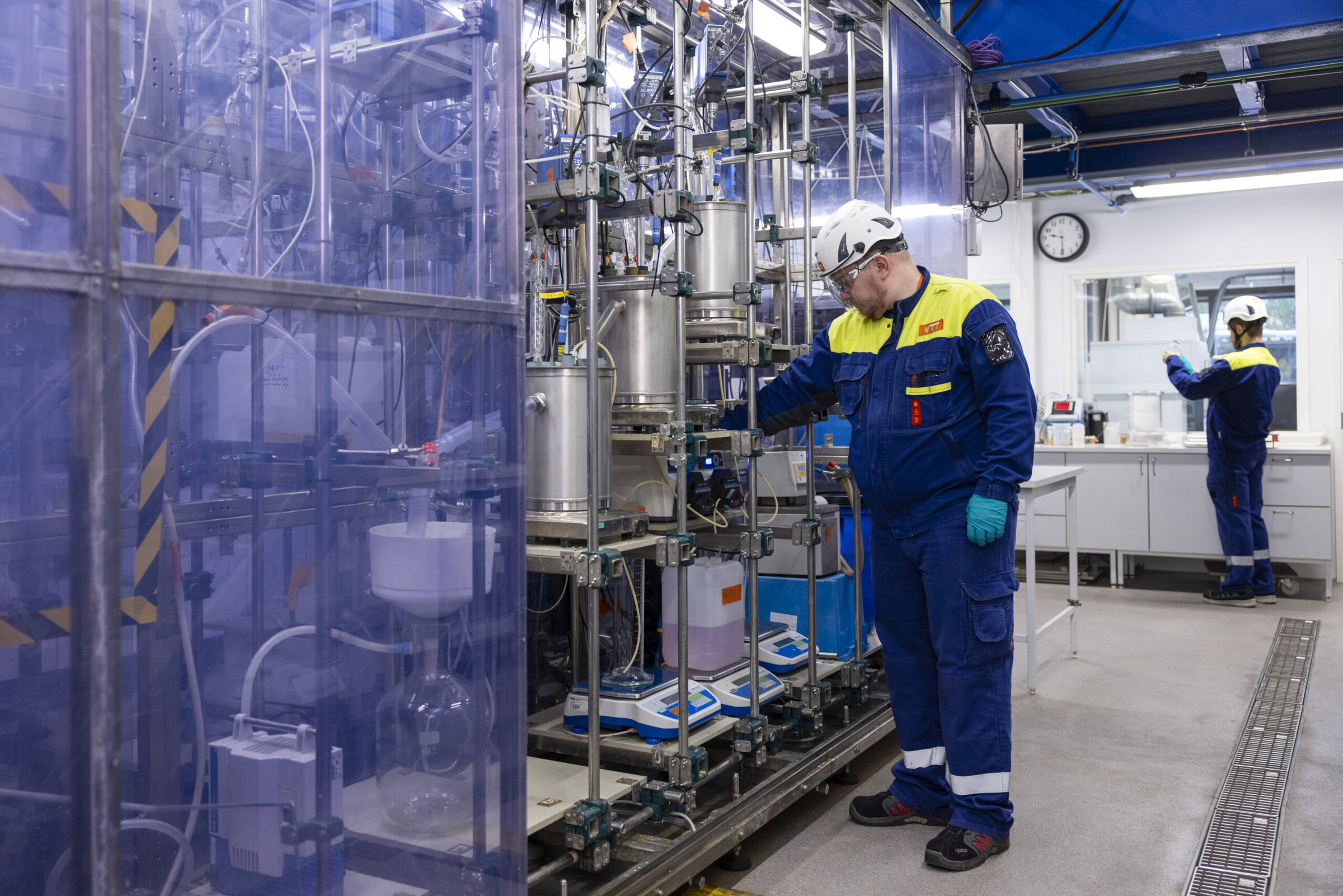 Two workers in protective gear operate machinery in a clean industrial lab environment.